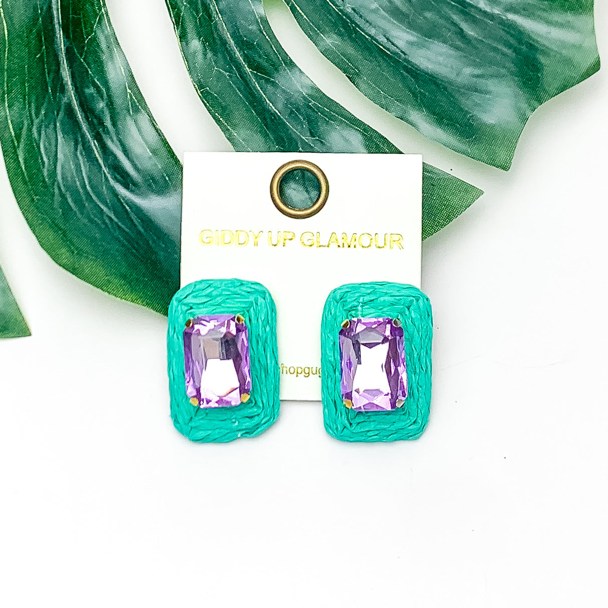 Truly Tropical Raffia Rectangle Earrings in turquoise green With Purple Crystal. Pictured on a white background with the earrings laying on a large leaf.