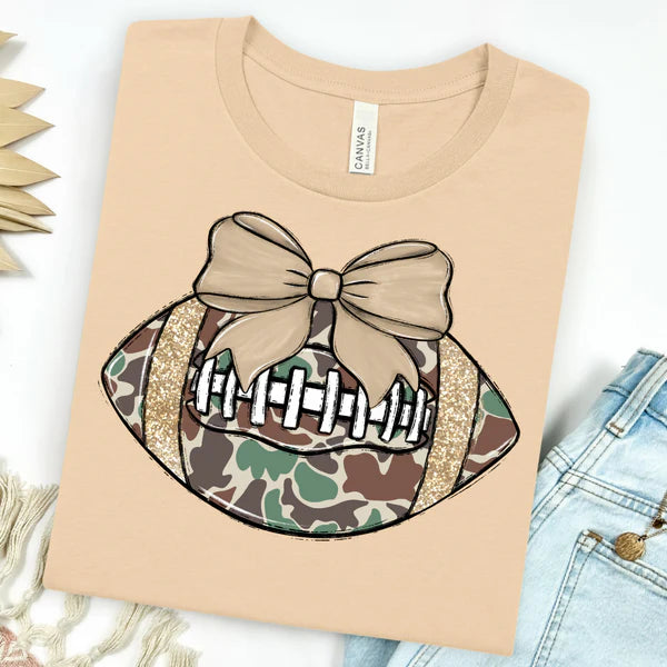 Cream short sleeve tee featuring a camo football graphic with a bow, combining sporty style and playful charm for casual wear.