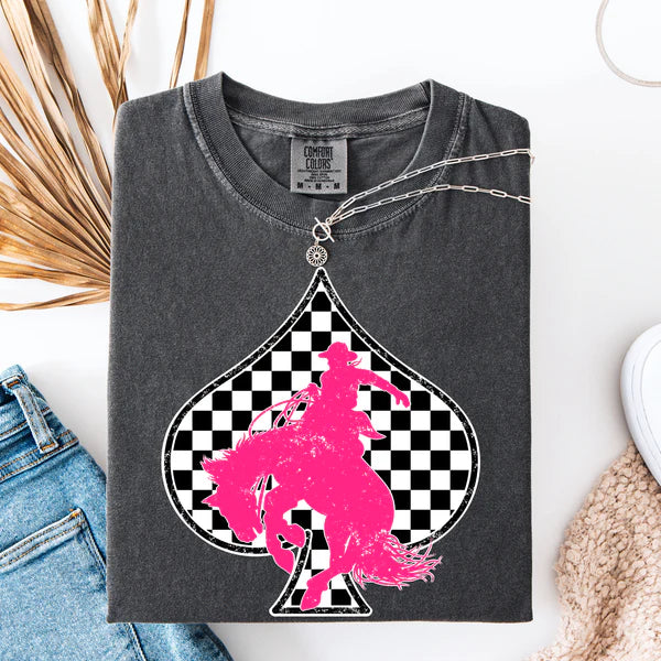 Folded dark gray t-shirt with pink graphic of a person on a horse in a checkered spade, displayed with jeans and decorative items.