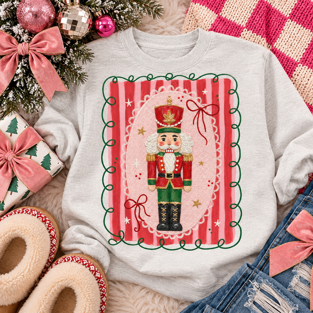 Classy Coquette Nutcracker Christmas graphic tee or sweatshirt on ash grey with a classic nutcracker in a pink striped frame with ribbon bows