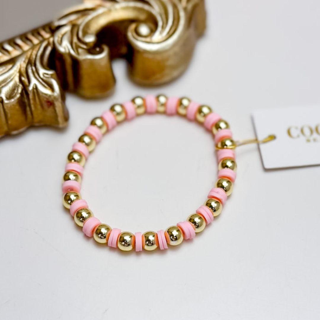 Bracelet with pink and gold beads on a white surface with a decorative gold object and a tag in the background.