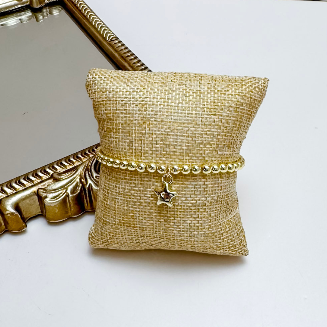 Gold bracelet with a star charm on a gold jewelry pillow next to a gold compact mirror.