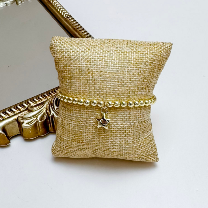 Gold bracelet with a star charm on a gold jewelry pillow next to a gold compact mirror.