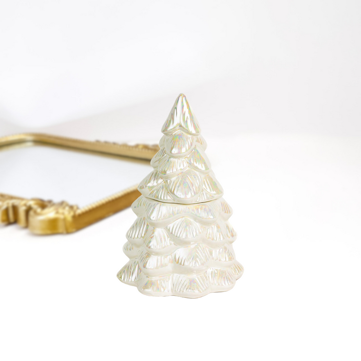A solid white background with a gold mirror and at the front a iridescent christmas tree candle by bridgewater candle company.