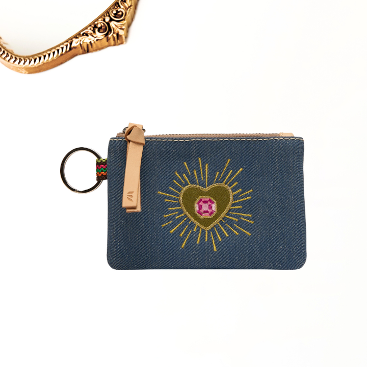 A small blue denim pouch with a tan zipper pull and key ring, featuring a gold heart with a pink gem motif on the front, shown on a white background with a cropped gold chain at the top left.