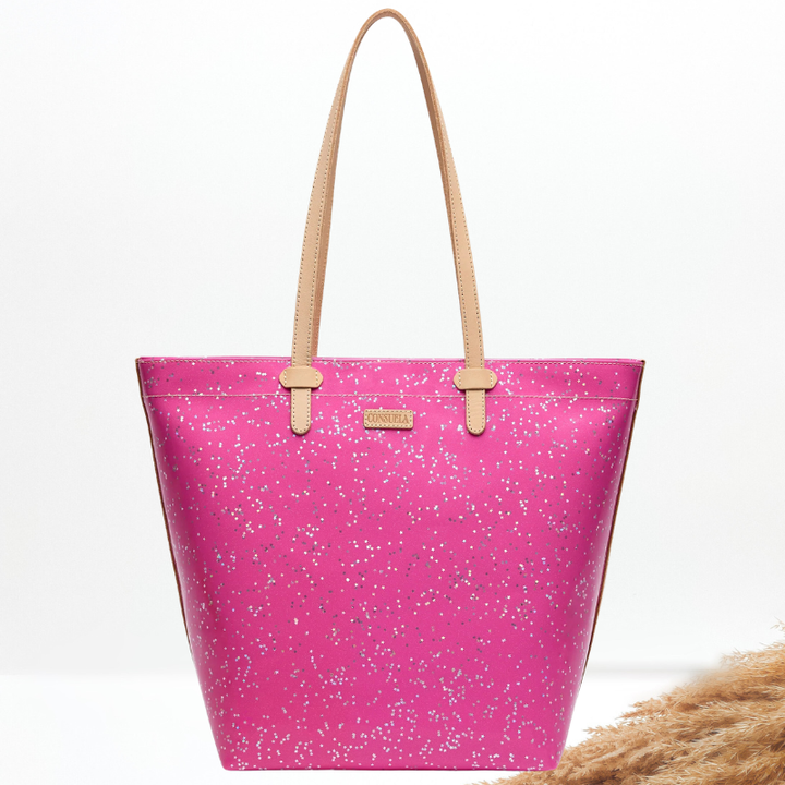 A bright pink tote bag with silver glitter speckles and light tan shoulder straps, shown on a clean white background with a decorative dried grass element in the lower right corner.