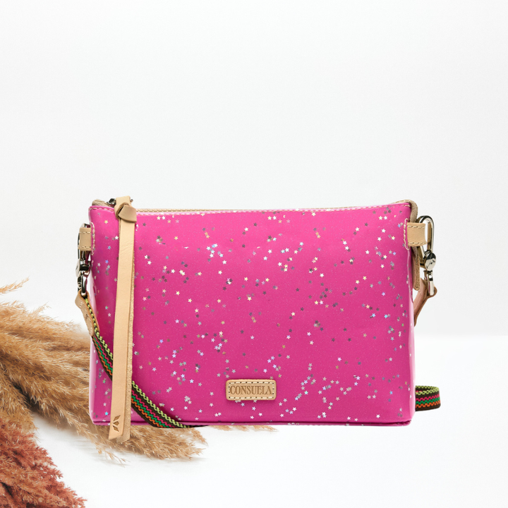 A bright pink crossbody bag with silver glitter speckles, tan leather trim, and a green and gold striped strap, displayed on a white background with dried pampas grass.