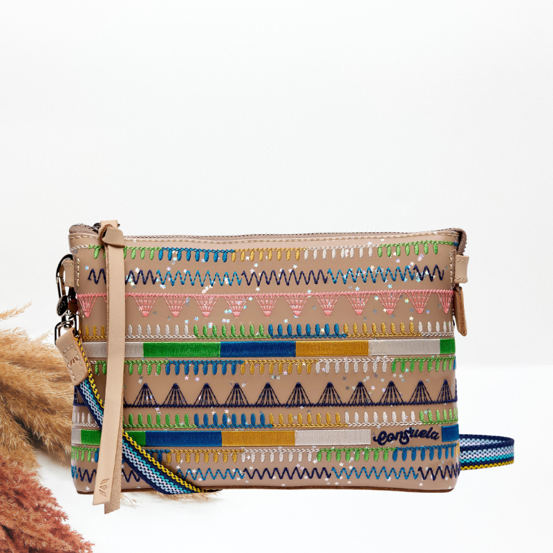 A rectangular woven clutch bag with a multicolor geometric pattern on a beige base, featuring a tan leather wristlet strap and a blue-and-white striped shoulder strap, photographed on a white background with dried grass as a prop.