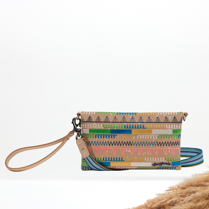 A multicolor geometric patterned wristlet clutch with a tan leather wrist strap and a blue patterned shoulder strap, displayed on a clean white background with a decorative beige pampas grass accent in the bottom right corner.