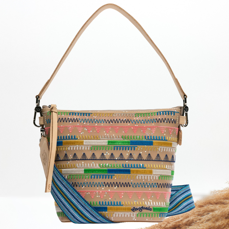 A multicolor patterned woven shoulder bag with a tan leather strap and a blue patterned detachable shoulder strap, displayed on a clean white background with a decorative brown pampas grass stem in the corner.