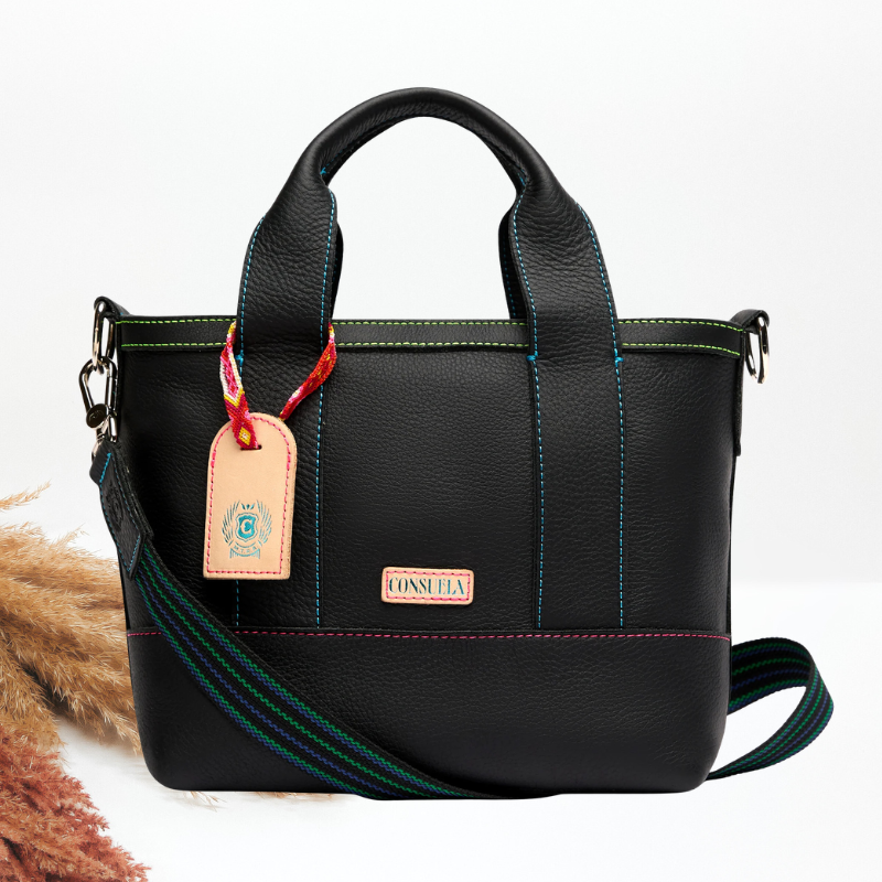 A black leather crossbody tote with multicolor topstitching, a striped woven shoulder strap, a leather luggage tag with a monogram charm, and a Consuela brand plate.