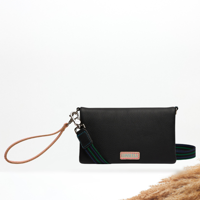 A black pebbled leather wristlet clutch with a tan leather wrist loop, a green and blue striped woven strap, and a small rectangular brand patch on the front, displayed on a white background with dried grass in the corner.