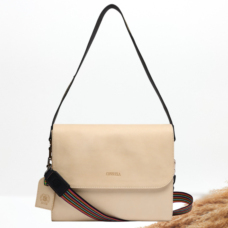 A beige leather crossbody bag with a black adjustable shoulder strap and a colorful striped woven crossbody strap, a leather luggage tag, and a Consuela brand imprint on the flap.
