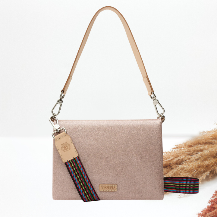 A champagne shimmer crossbody bag with a tan leather shoulder strap, a multicolor striped guitar strap and a leather logo tag, styled with dried pampas grass on a white background.