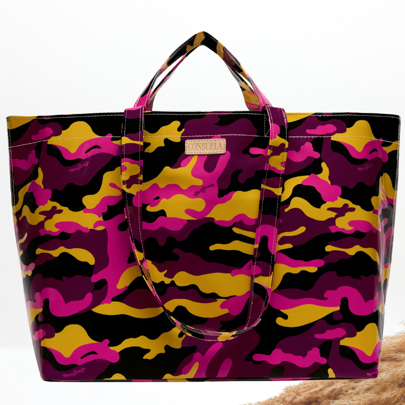 A large tote bag featuring a bold camouflage pattern in black, magenta pink, and mustard yellow. The bag has dual top handles, a small tan leather brand patch on the front, and is photographed against a clean white background.