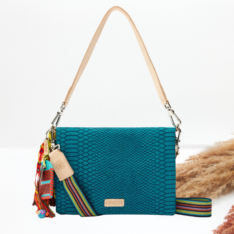 A teal blue-green snakeskin-pattern shoulder bag with a beige leather strap, colorful tassels, and a striped woven strap detail, shown on a white background with dried grass props.