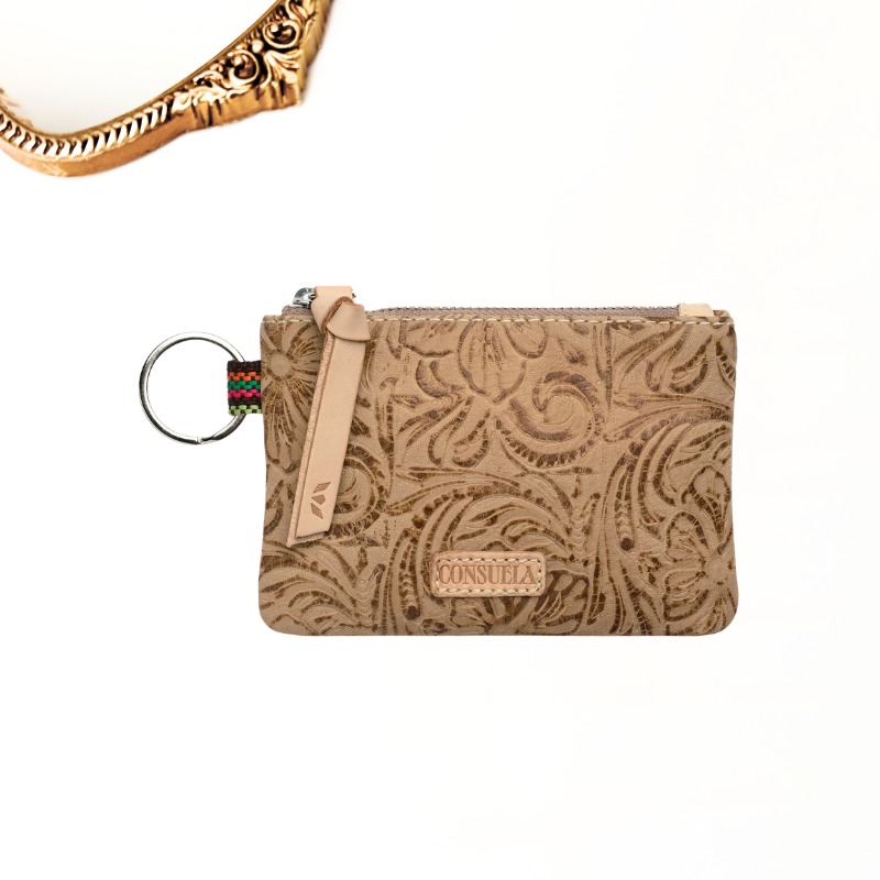 A tan tooled leather pouch with an embossed paisley pattern, featuring a matching leather zipper pull, a small woven accent, and a metal key ring. A brand label on the front reads 'CONSUELA'.