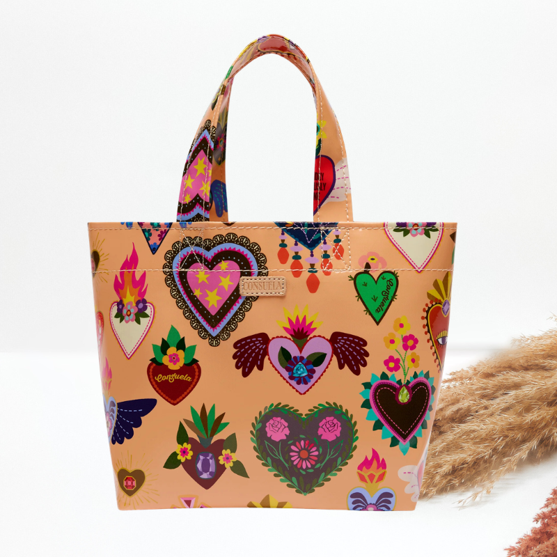 A peach-colored tote bag with a vibrant, multicolored print of Sacred Heart of Jesus motifs, including hearts, flowers, and angels, shown on a white background with dried grass as a prop.