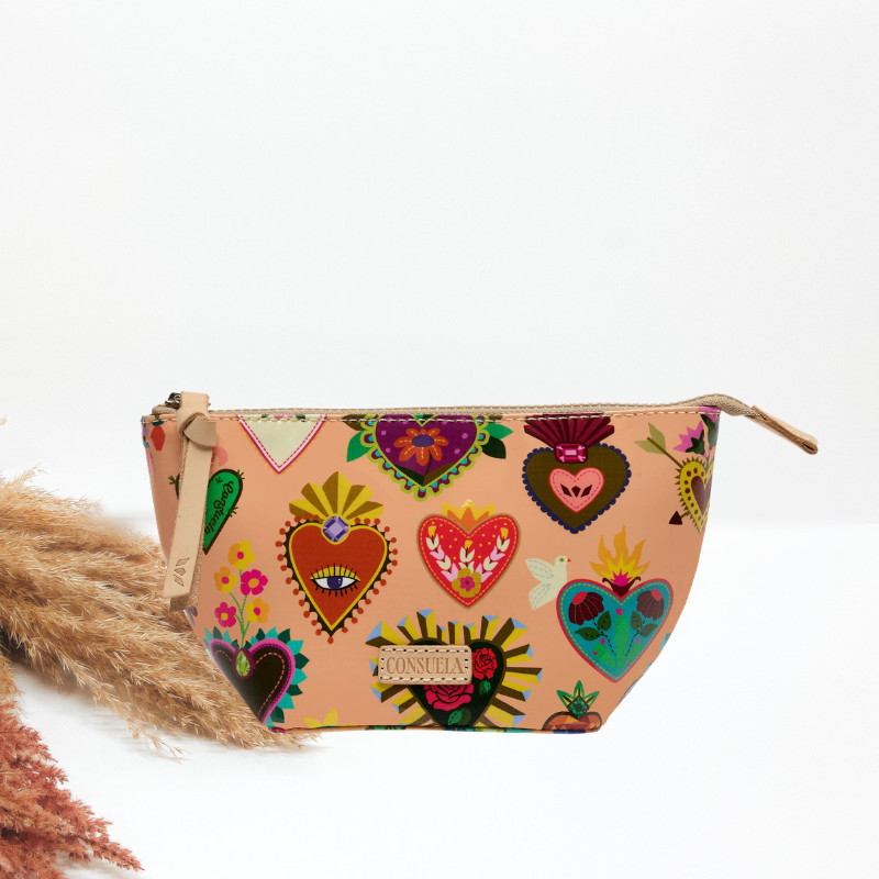 A small zippered pouch with a beige base and a colorful print of Sacred Heart of Jesus motifs, including hearts with eyes, flames, and floral details. The pouch is styled on a white background with dried pampas grass on the left.