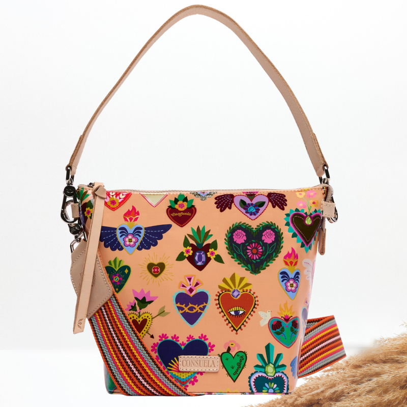 A Consuela shoulder bag with a peach-colored base covered in colorful embroidered hearts and floral motifs, featuring a tan leather strap and a striped woven crossbody strap.