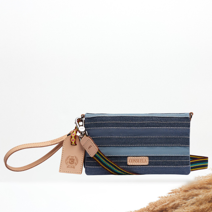 A Consuela wristlet clutch with a blue and navy striped canvas body, a tan leather wrist strap and hangtag, and a multicolor woven shoulder strap.