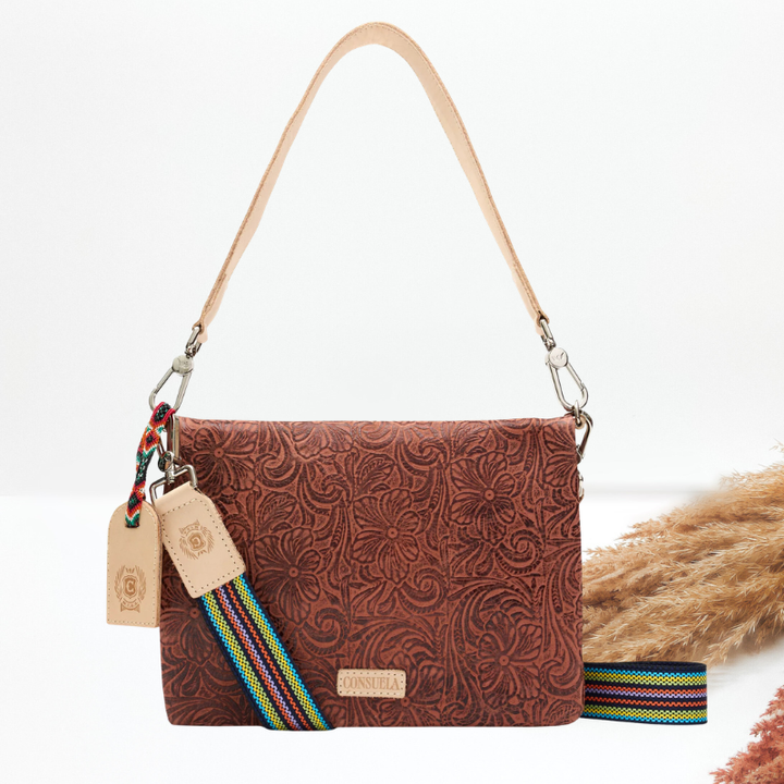 A brown tooled leather crossbody bag with a tan leather shoulder strap, a multicolor striped guitar strap, and a leather logo tag, styled with dried pampas grass on a white background.