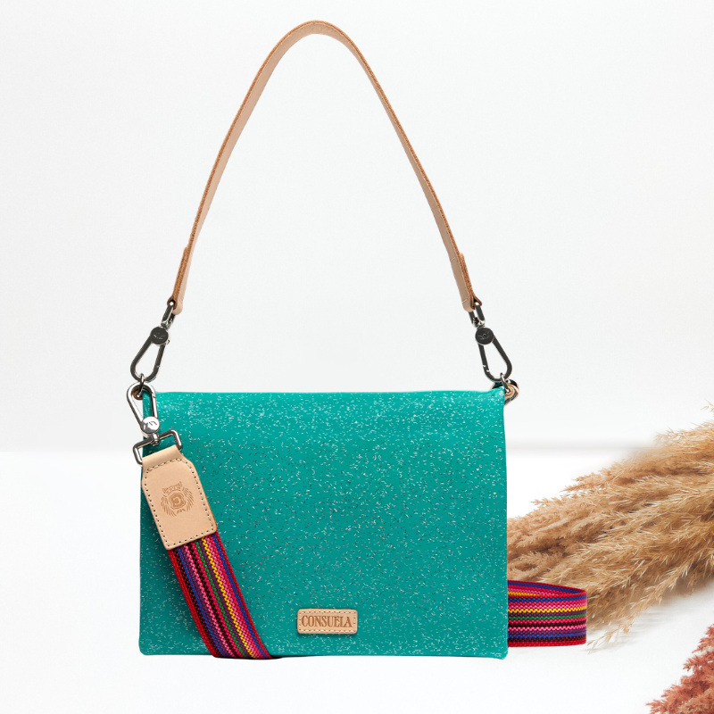 A turquoise glitter shoulder bag with a tan leather strap and a multicolor woven accent strap, displayed on a white background with dried pampas grass.