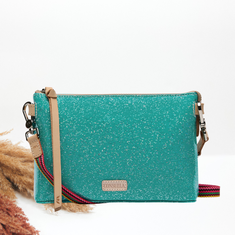 A turquoise glitter crossbody bag with a beige leather strap and a multicolor woven strap detail, shown on a white background with decorative dried grass on the left.