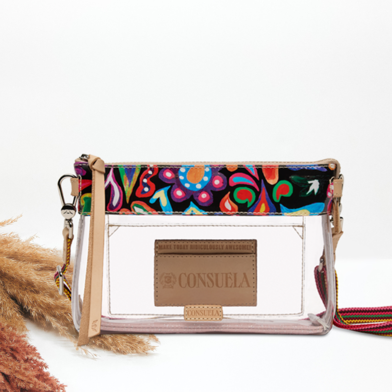 A clear plastic crossbody clutch bag with a colorful patterned top band, beige leather trim, and a tan brand patch reading CONSUELA. The bag is photographed on a white studio background with dried grass stems at the lower left.