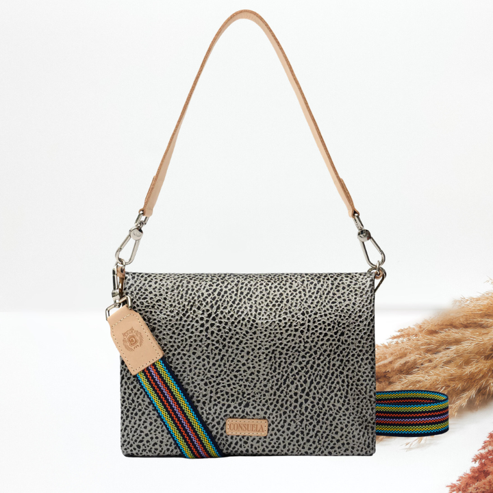 A gray and black speckled hair on hide crossbody bag with a tan leather shoulder strap, a multicolor striped guitar strap and a leather logo tag, styled with dried pampas grass on a white background.