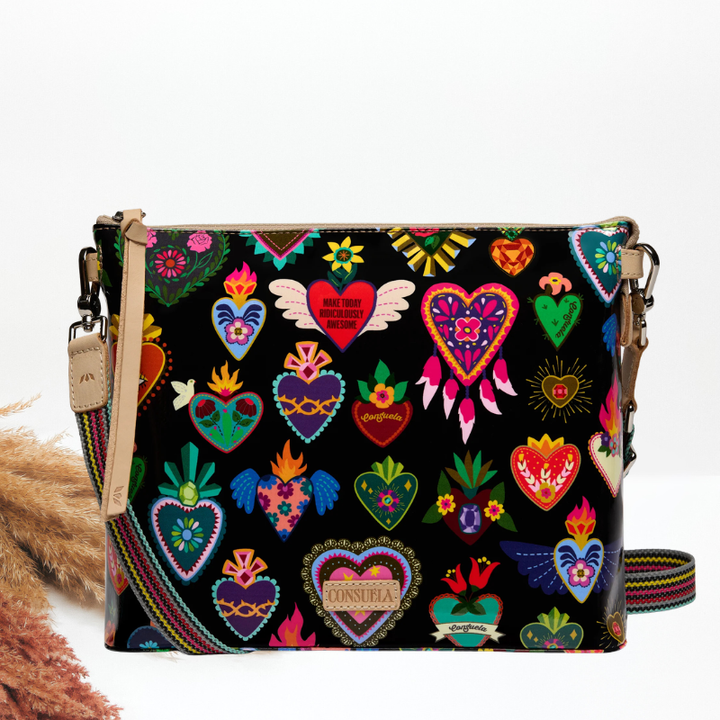 A black crossbody bag with a colorful embroidered-style pattern of hearts, wings, flowers, and folk-art motifs, a tan leather strap, and a leather &