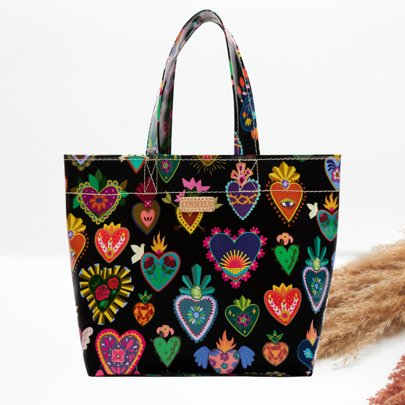 A black tote bag with colorful embroidered-style Sacred Heart motifs and dual shoulder straps, photographed against a clean white background with a small decorative dried plant prop at the lower right.