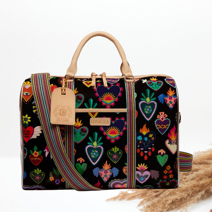 A black duffel bag with a colorful folk-art heart print, tan leather handles, a striped woven crossbody strap, a leather luggage tag with a monogram charm, and a Consuela brand plate.