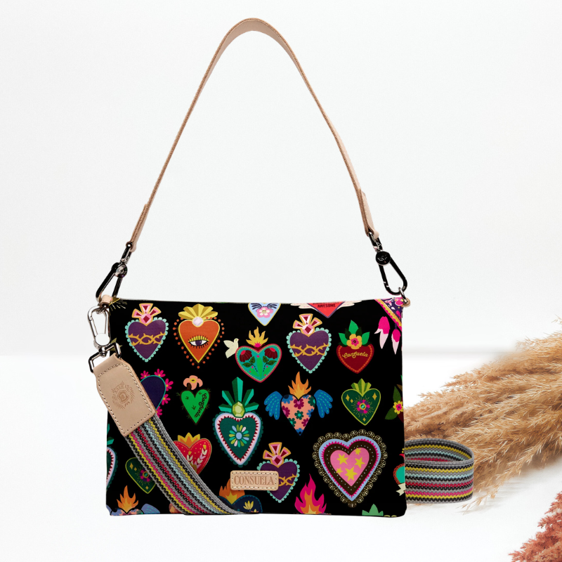 A black shoulder bag with a colorful embroidered Sacred Heart pattern, featuring a tan leather strap and a matching woven crossbody strap, displayed on a white background with dried grass.