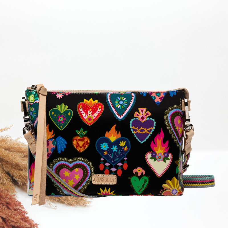 A black crossbody bag with colorful embroidered Sacred Heart designs, a tan leather strap, and a woven shoulder strap, shown on a white background.