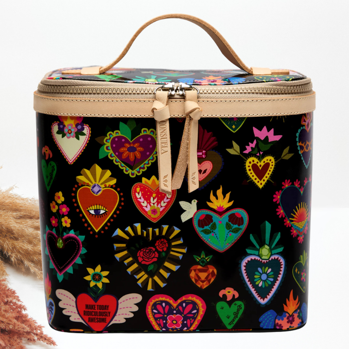 A cylindrical cosmetic bag with a black background covered in colorful heart illustrations, featuring a tan leather handle and zipper trim, photographed on a white background with dried grass as a prop.