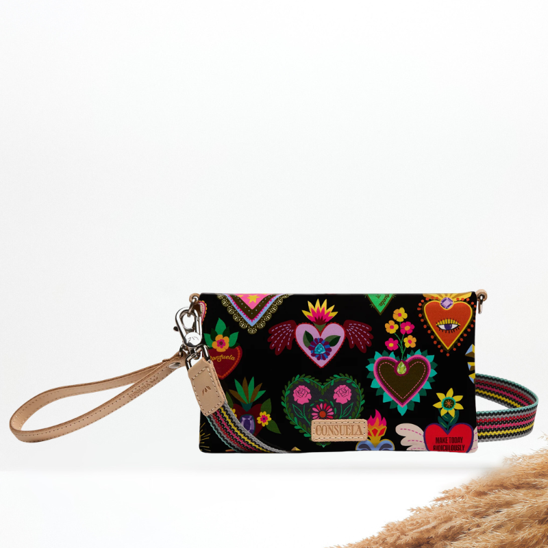 A black wristlet clutch with a colorful folk-art heart pattern, featuring a tan leather wrist strap and a multicolor woven strap, set against a white background with a decorative straw prop in the corner.