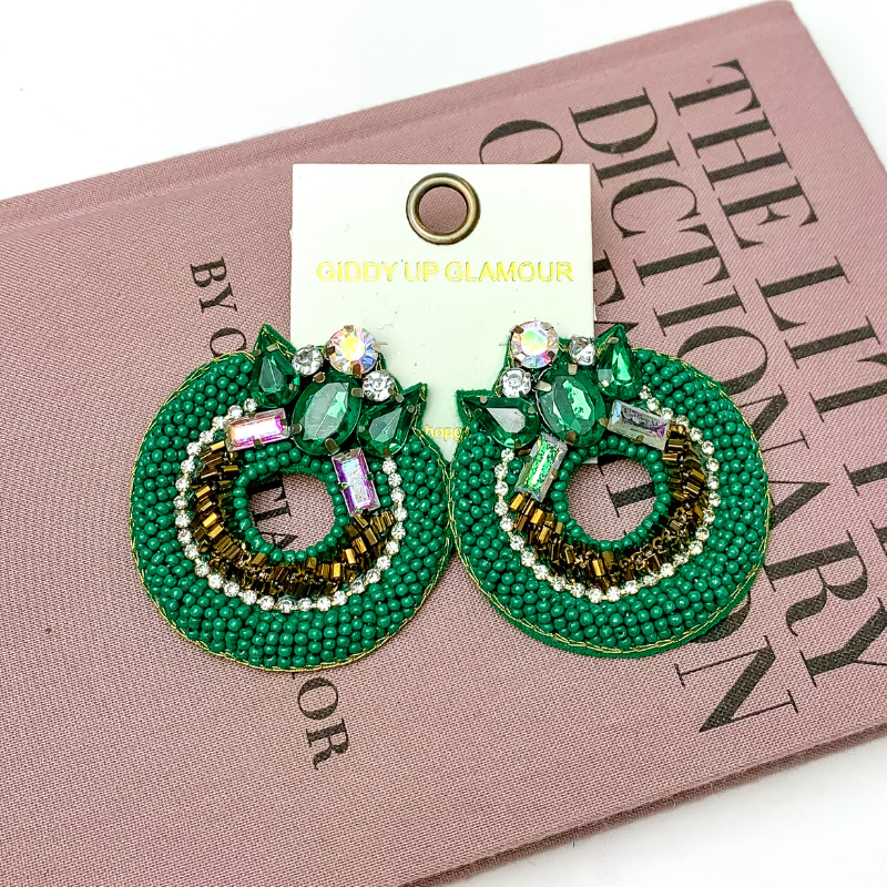 Pictured is hoop beaded green and bronze earrings with a jeweled detailing at the top. These earrings are pictured on a white background with a pink fashion dictionary.