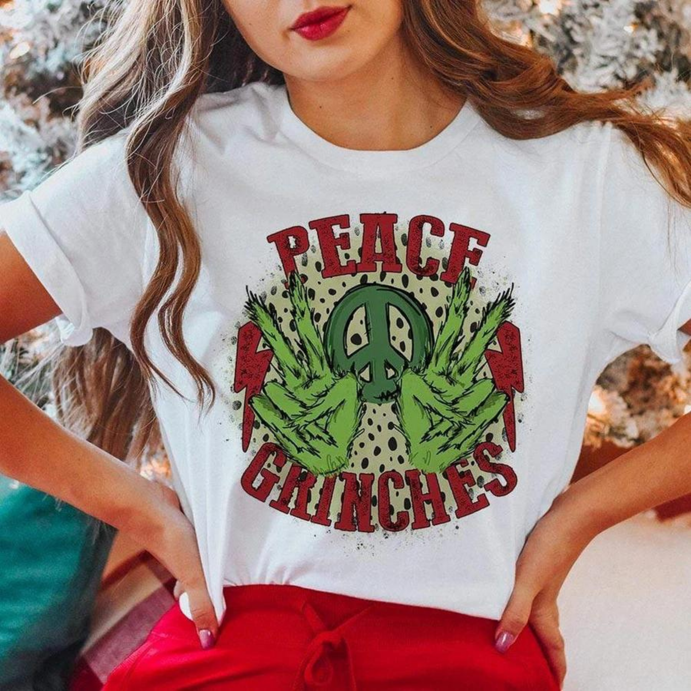 This white tee includes a crew neckline, short sleeves, and a cute Christmas graphic with a leopard background that says "Peace Grinches" in red font, two Grinch hands throwing up the peace sign, a green peace sign between the hands, and two red lightning bolts on the outside of the Grinch hands. The model has this graphic tee styled with rolled sleeves and a pair of red pants. 