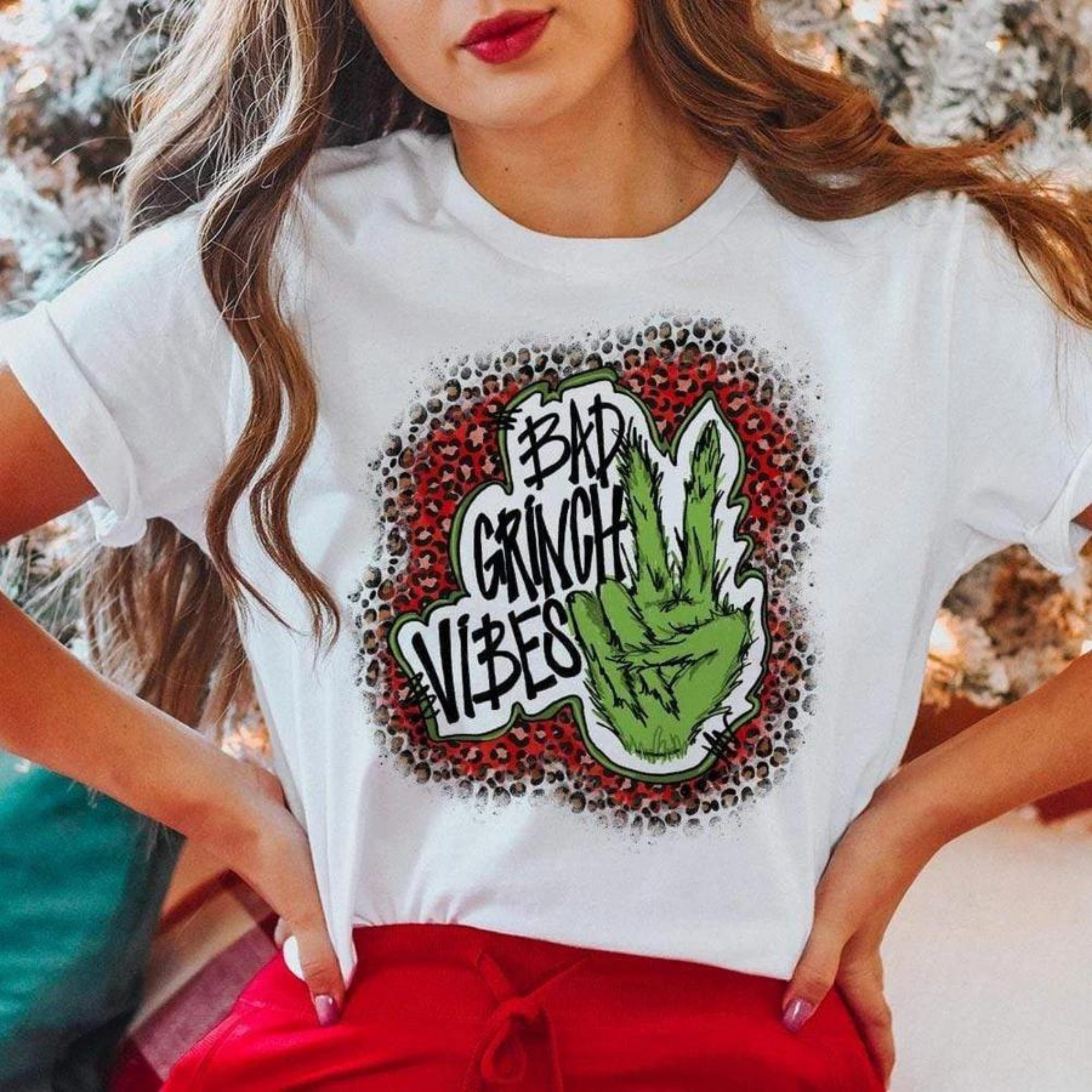 Model is wearing a white tee shirt that has a red leopard graphic with a grinch hand throwing a peace sign. The graphic says "Bad Grinch Vibes". This graphic tee is styled with rolled sleeves and a pair of red pants. 