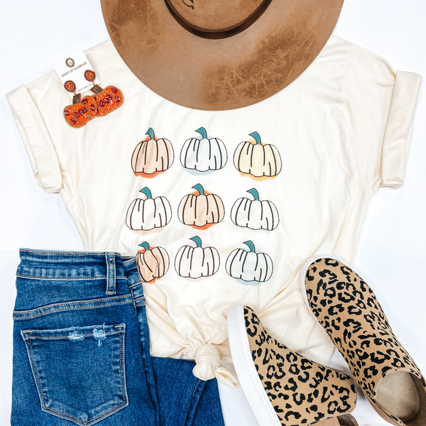 White tee with nine pumpkins printed on it. Pumpkins are white or light orange in color. Tee is styled with items that complete the outfit - a tan Charlie One Horse Hat, pumpkin earrings, leopard print sneakers and medium wash jeans. 