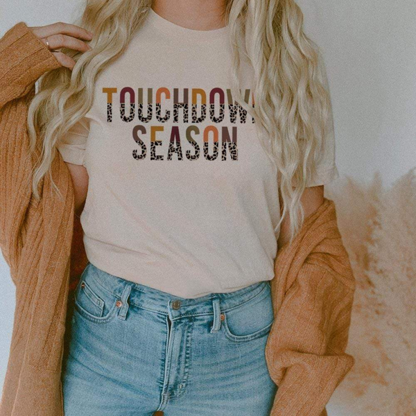 Model is wearing a cream color short sleeve crewneck featuring a half multicolor half leopard print graphic that says "Touchdown season"