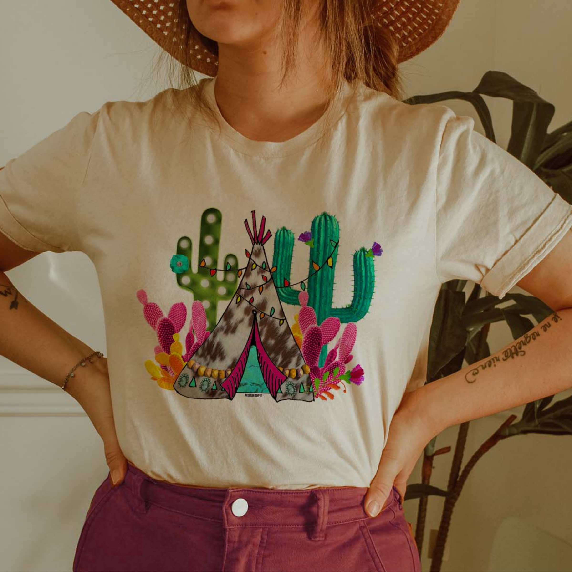 This cream tee includes a crew neckline, short sleeves, and a teepee and cactus graphic in bright, fun colors. The model is shown here wearing the tee with rolled sleeves, a light straw hat, silver bracelet, and fuchsia pair of denim jeans. 