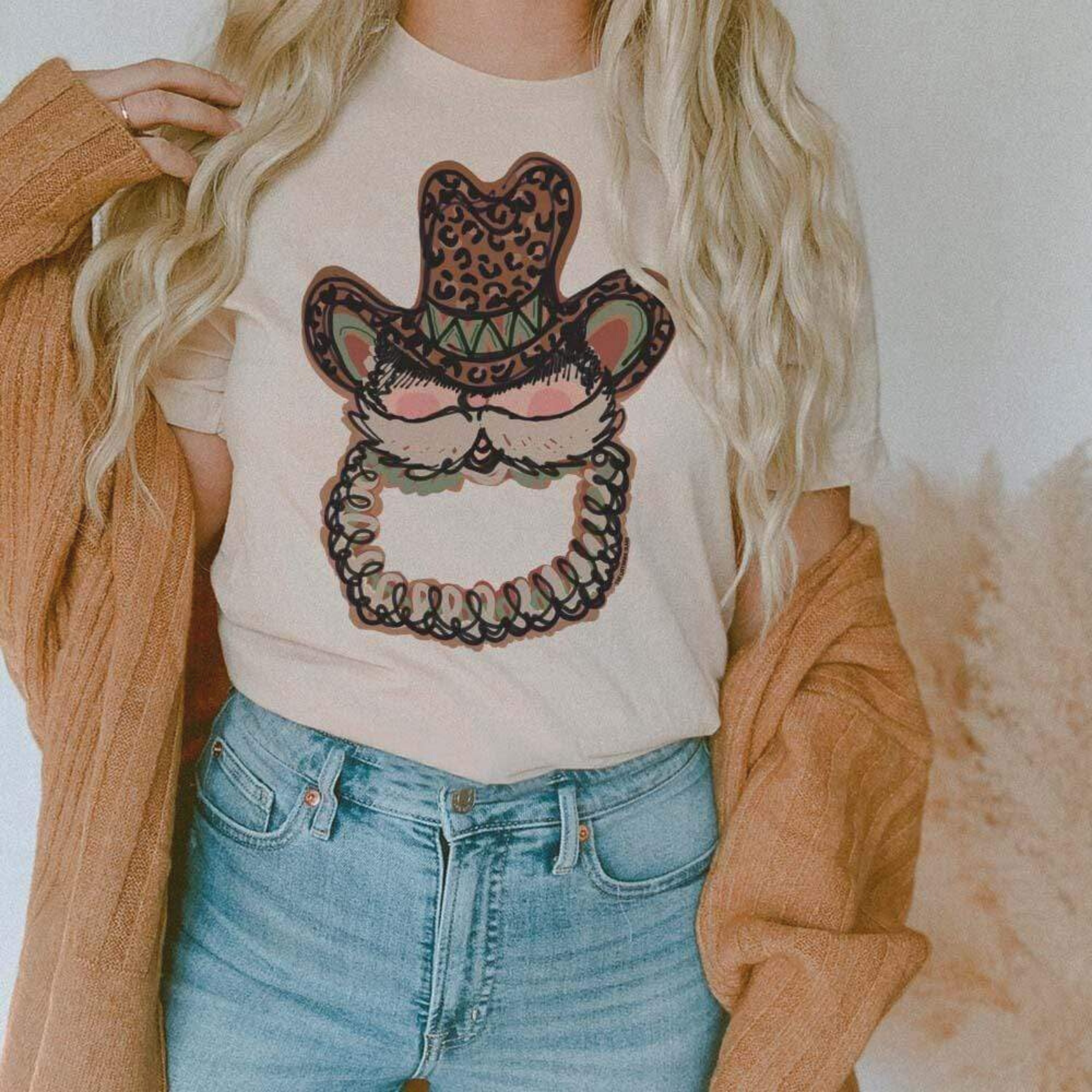This cream tee includes a crew neckline, short sleeves, and a Santa Clause graphic with him wearing a leopard print cowboy hat. The model has this tee styled with rolled sleeves, a camel cardigan, and a light wash pair of denim jeans. 