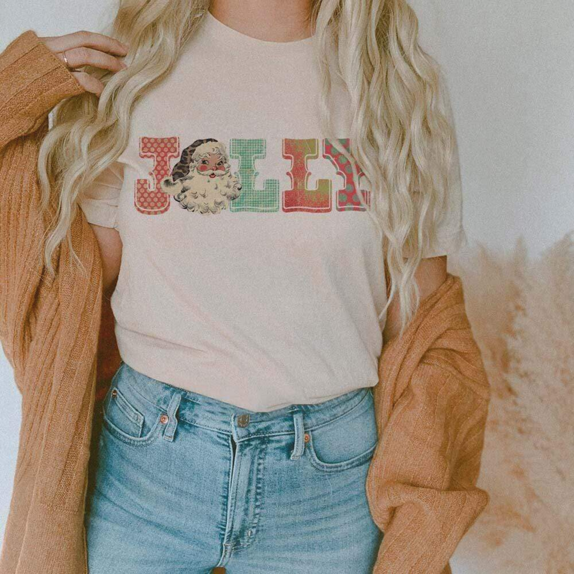 This cream tee includes a crew neckline, short sleeves, and a graphic that says "Jolly" in a cute, festive font with a Santa Clause graphic serving as the "O" in the word "JOLLY". The model has this tee shown with rolled sleeves, a camel cardigan, and a light wash pair of denim jeans. 