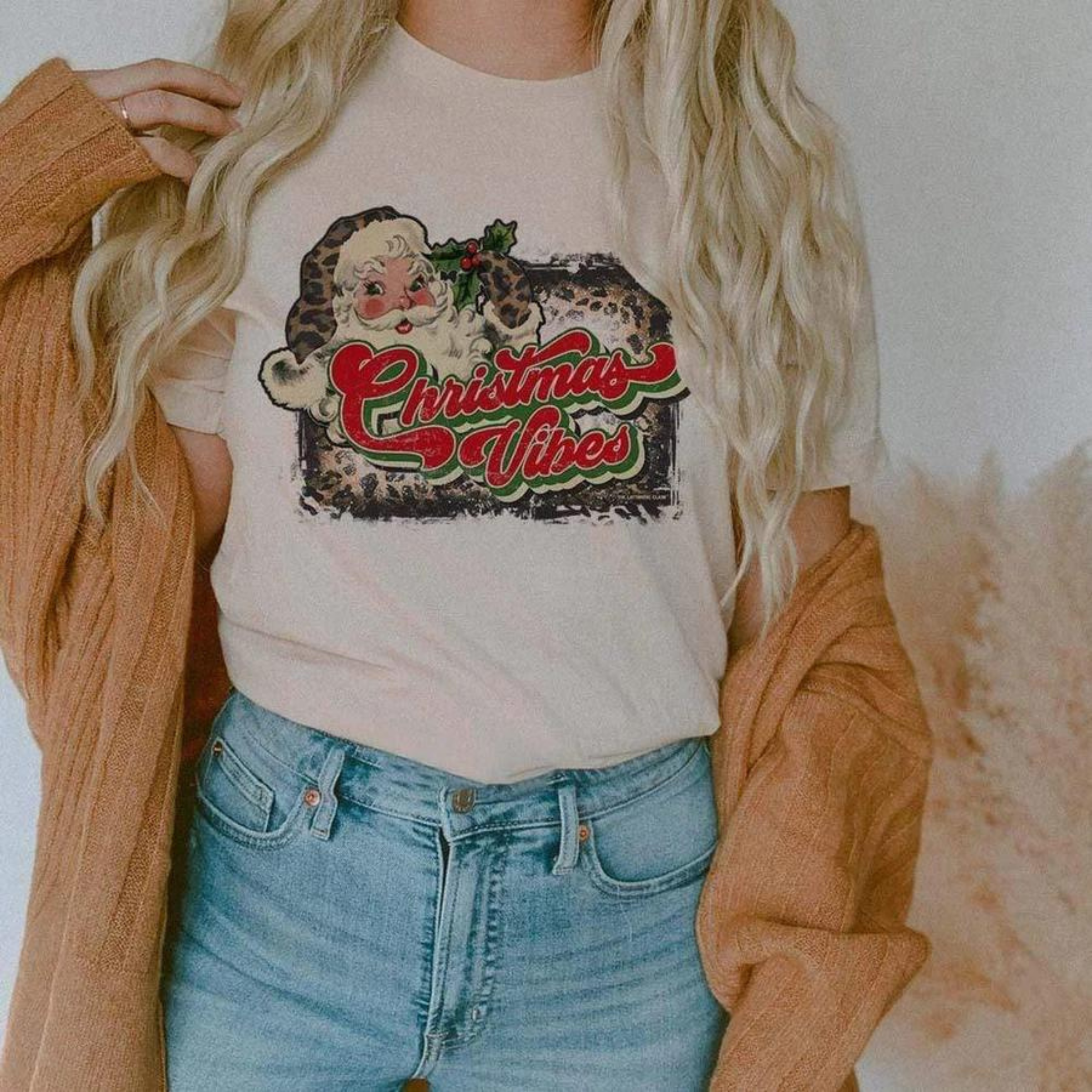 This cream tee includes a crew neckline, short sleeves, and a cute Christmas graphic that says "Christmas Vibes" in a red and green cursive font. There is a Santa Clause wearing a leopard hat and glove, as well as a bleached leopard background behind the saying and Mr. Clause. The model has this graphic tee styled with rolled sleeves, a camel cardigan, and a light wash pair of denim jeans. 