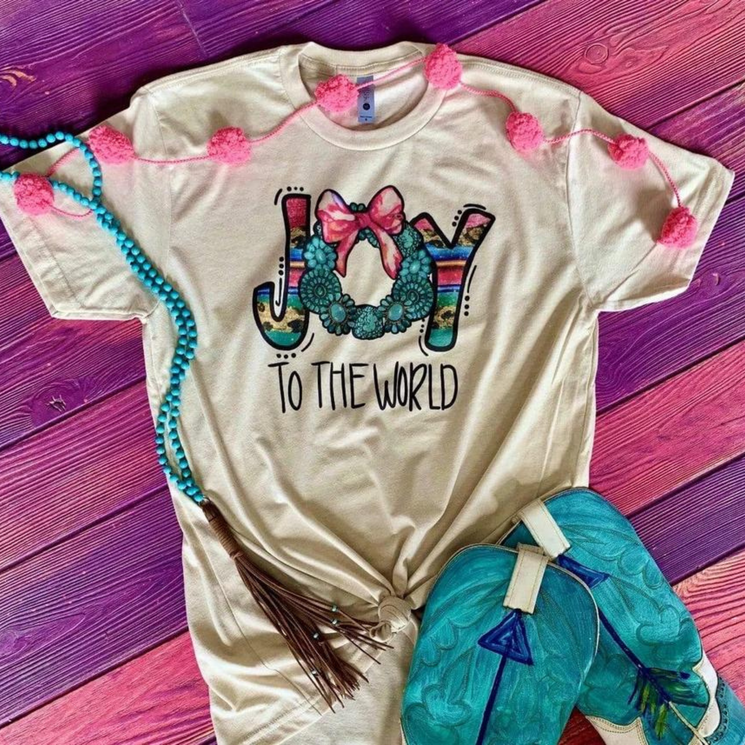 This cream tee includes a crew neckline, short sleeves, and a cute Christmas graphic with the saying "Joy to the World" with the word "JOY" in an uppercase font with serape inside of the J + Y, and the O with turquoise pieces and a pink bow. The words "TO THE WORLD" are uppercase in a handwritten font under the word "JOY". This tee is shown here as a flat lay paired with turquoise cowboy boots and a turquoise necklace with a brown tassel. 