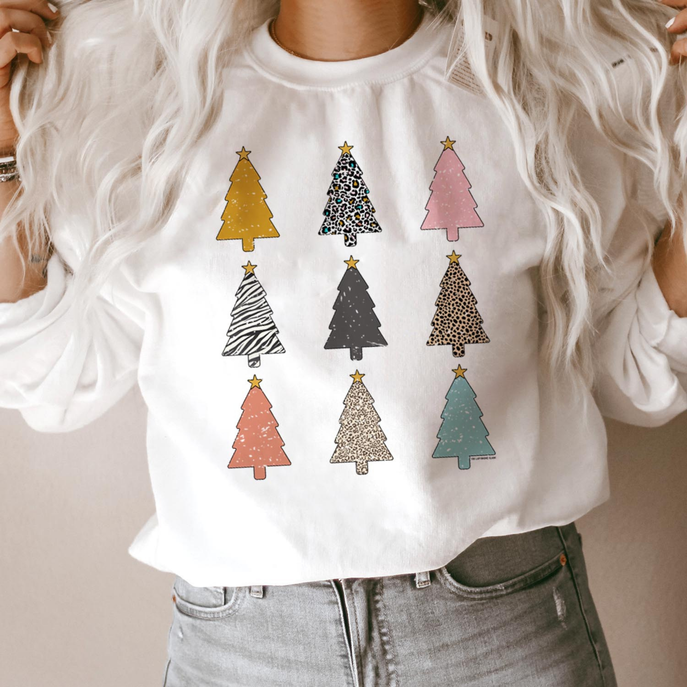 This white pullover includes a crew neckline, long sleeves, and three rows of three multi print Christmas tree collage graphic. This sweatshirt is modeled with rolled sleeves and a light wash pair of denim jeans. 
