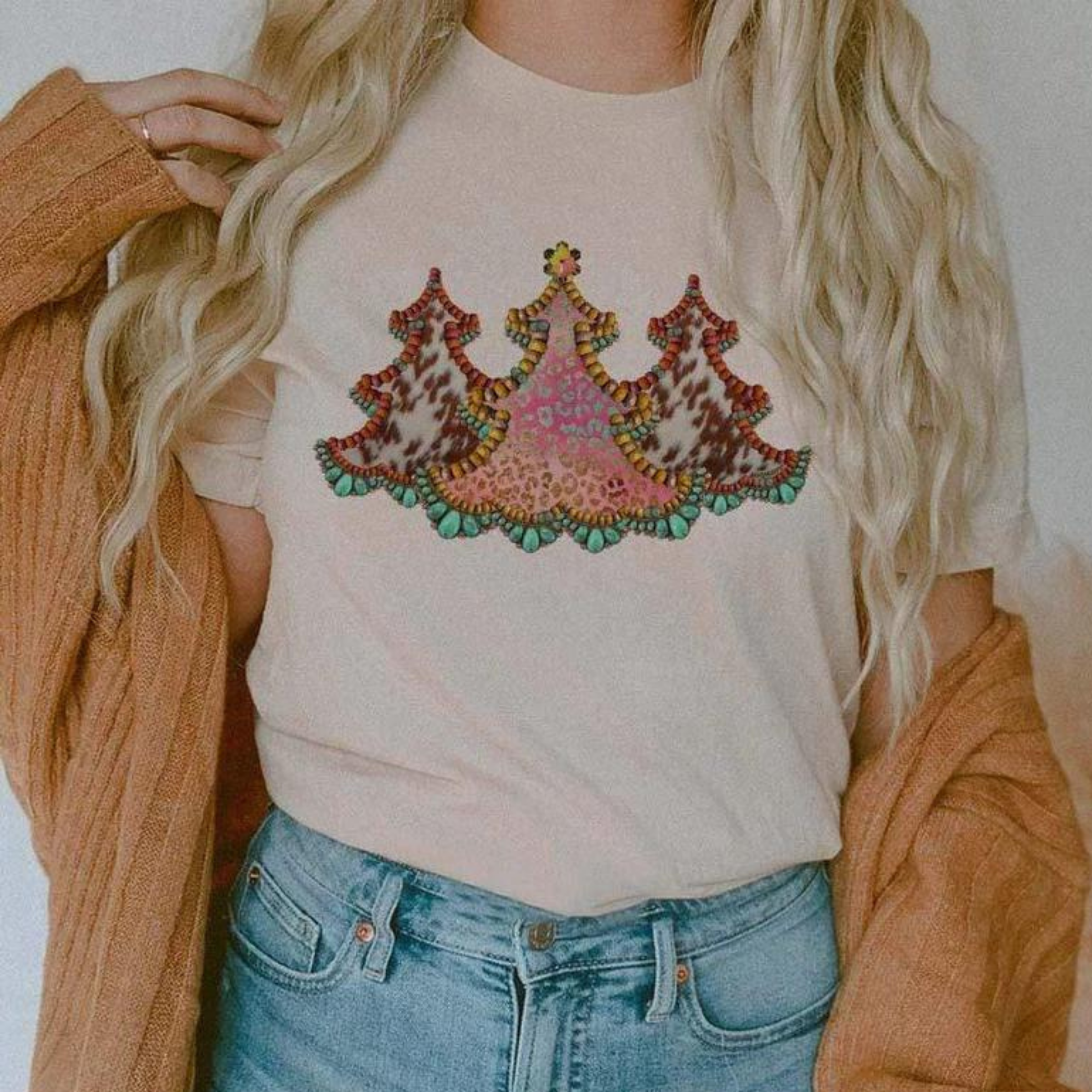 This cream tee includes a crew neckline, short sleeves, and a cute Christmas graphic with three cowprint Christmas trees side by side. The model has this graphic tee styled with rolled sleeves, a camel cardigan, and a pair of light wash denim jeans. 
