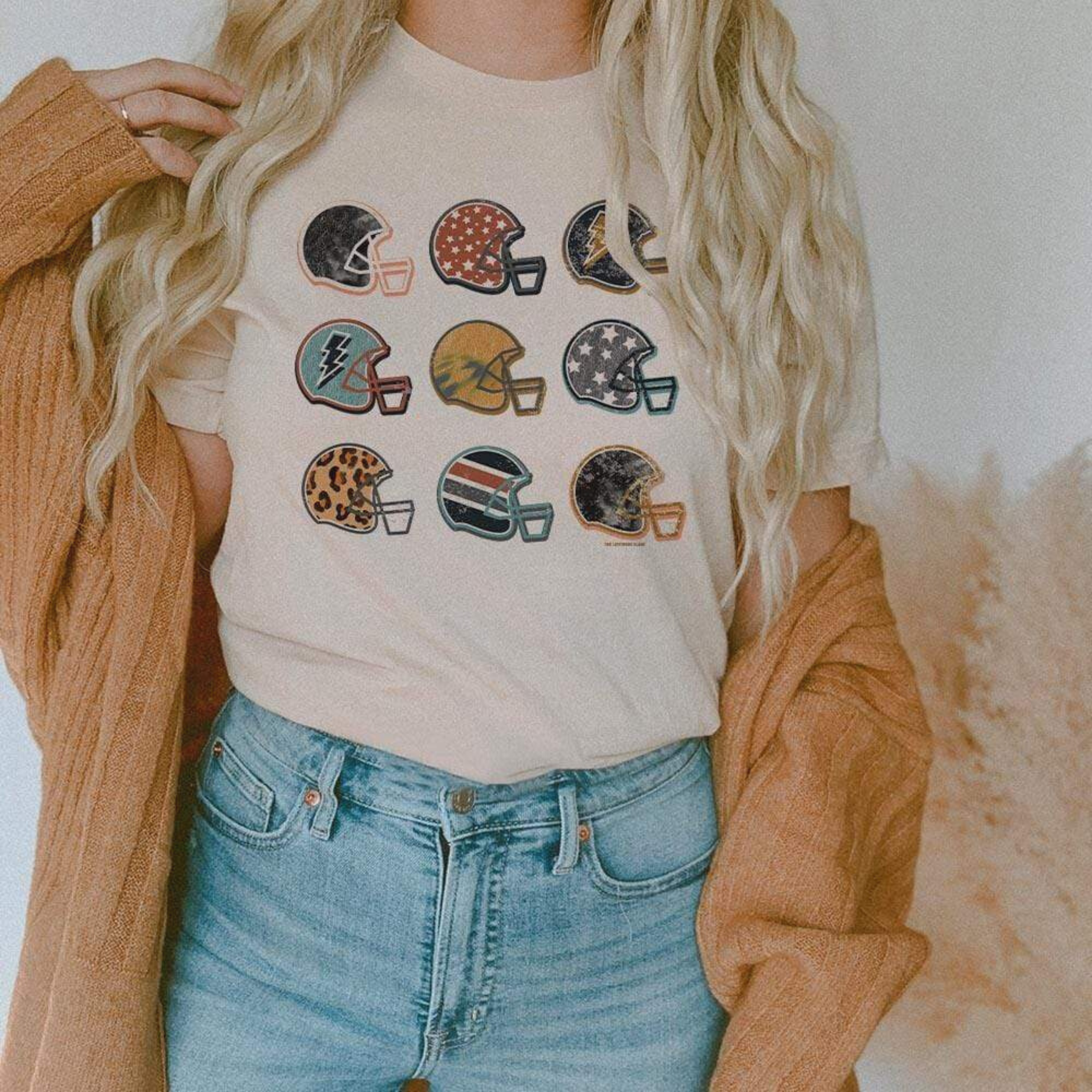 Model is wearing a cream short sleeve crewneck tee featuring a graphic of 9 football helmets, each with their unique pattern.